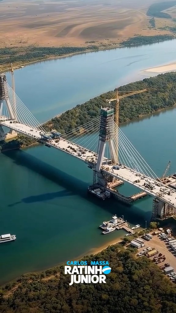 Vista aérea de uma ponte em construção sobre um rio, com guindastes e veículos em atividade nas proximidades.