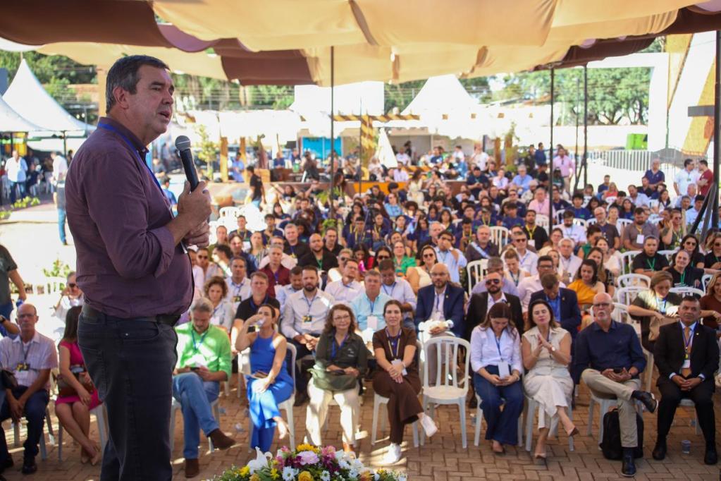Um palestrante em pé, segurando um microfone, fala para uma multidão em um evento ao ar livre. O público é composto por diversas pessoas sentadas olhando atentamente. Há tendas e decoração ao fundo.
