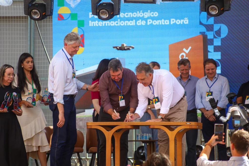 Evento de assinatura formal em Ponta Porã, com várias pessoas em pé e uma mesa na frente onde dois homens assinam documentos. Ao fundo, um telão com o nome do evento.