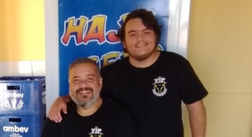 Dois homens sorrindo, um sentado e outro em pé ao lado, ambos vestindo camisetas pretas. Ao fundo, uma parede com uma faixa colorida que diz 'HÁJ CERTO'.